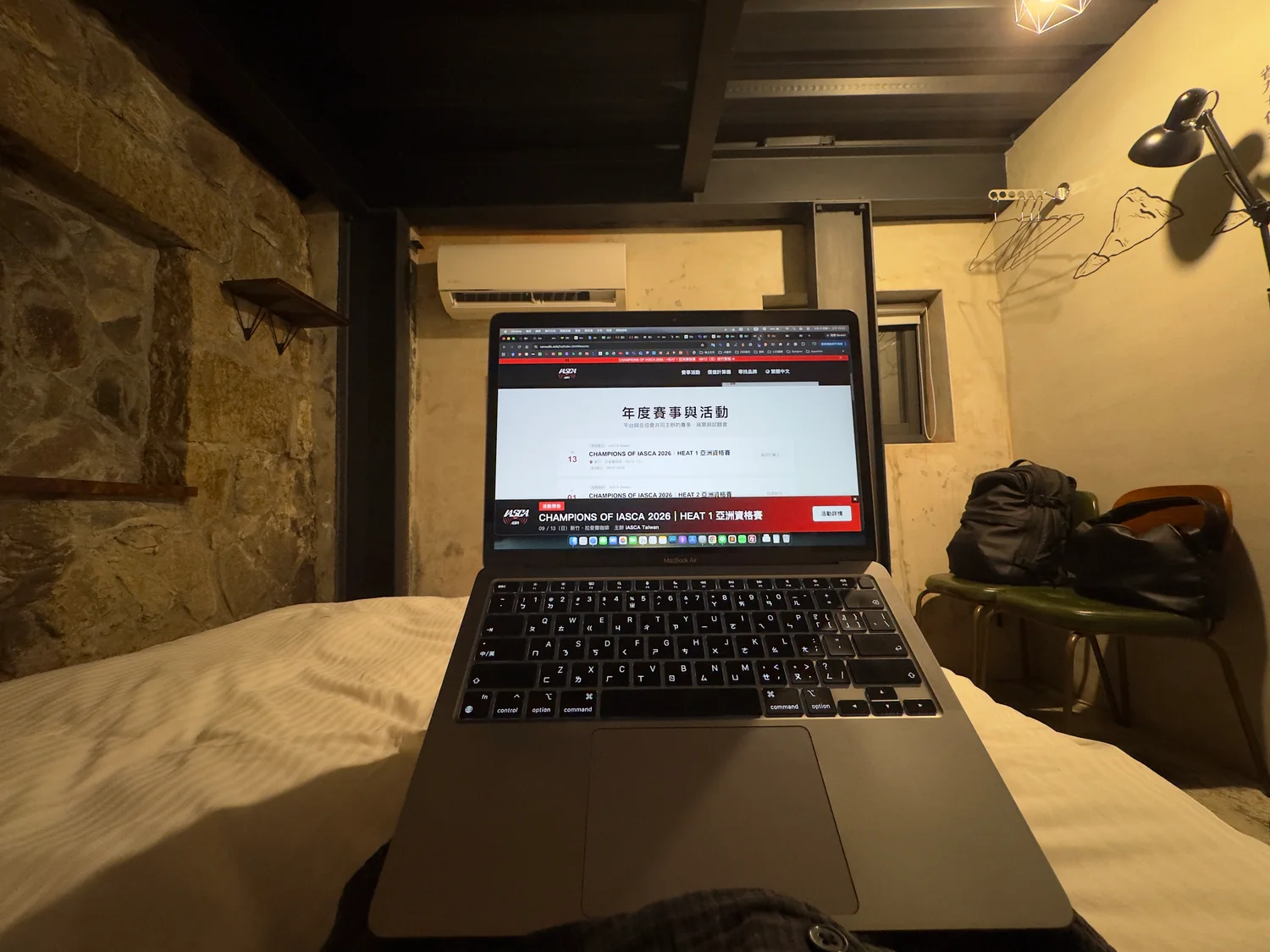從床上視角拍攝的 MacBook,電腦放在身前,背景是房內石牆、冷氣與掛衣處