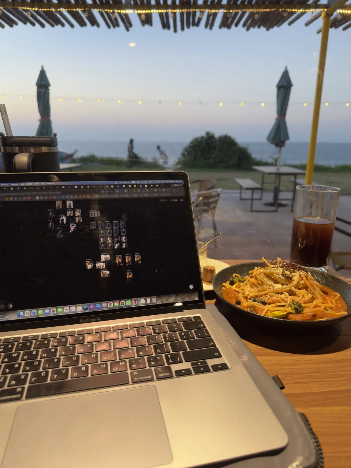 傍晚的戶外座位亮起串燈,桌上是 MacBook、義大利麵與冰美式